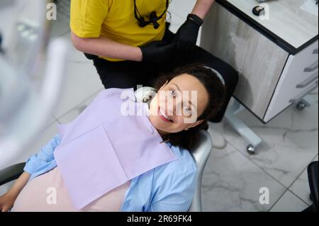 Porträt einer jungen schwangeren Frau im Zahnarztstuhl, lächelnd in die Kamera während einer Zahnarztuntersuchung. Blick von oben Stockfoto