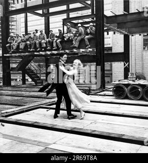 Vereinigte Staaten: c. 1959. Ein attraktives junges Paar in formeller Abendgarderobe tanzt auf einer Baustelle, während eine Reihe von Arbeitern auf einer Stahlbalkenwache ihr Mittagessen essen. Stockfoto