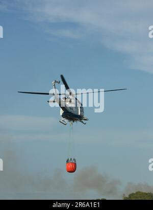 Ein Helikopter fliegt mit dem Bambi-Eimer-System zusammen, um ein Feuer in der Nähe zu löschen, das Wasser aus einem Kanal transportiert Stockfoto