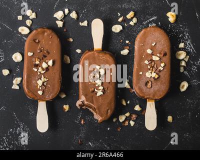 Milchschokolade Eis am Stiel mit Haselnüssen. Nahaufnahme. Eis am Stiel mit Schokolade, Stäbchen und schwarzem Steinhintergrund. Draufsicht. Schokolade Stockfoto