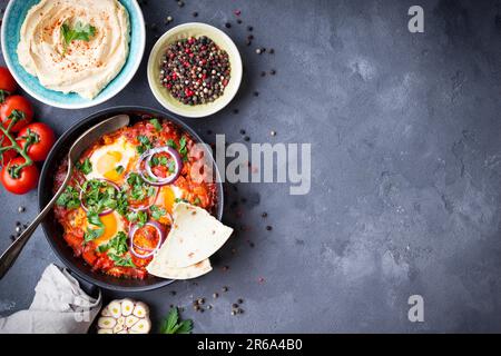 Shakshuka mit Pita-Brot in der Pfanne, Hummus in der Schüssel auf rustikalem Hintergrund. Traditionelle Gerichte aus dem Mittleren Osten. Spiegeleier mit Gemüse. Draufsicht. Leerzeichen Stockfoto