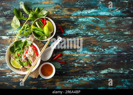 Traditionelle vietnamesische Suppe pho bo mit Kräutern, Fleisch, Reis, Nudeln, Suppe. Pho Bo in der Schüssel mit Stäbchen, Löffel. Platz für Text. Ansicht von oben. Asiatische Suppe Stockfoto