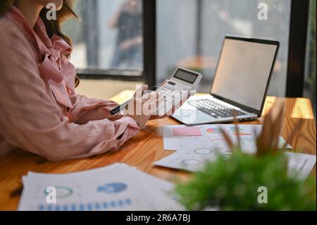 Zugeschnittenes Bild einer schönen asiatischen Geschäftsfrau oder Buchhalterin in einem rosafarbenen Kleid mit einem Taschenrechner und Analyse von Finanzdaten an ihrem Schreibtisch. Stockfoto