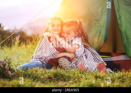 Zwei kleine umarmende Mädchen Schwestern sitzen auf dem grünen Gras neben dem Zelteingang und lächeln fröhlich. Sorglose Kindheit, Familienwerte und Outdoor Stockfoto