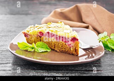 Ein Stück süßen Kuchen mit Himbeeren, Butterbröckeln und Mandelflocken auf einem Teller, Minze und Serviette auf Holzbretthintergrund Stockfoto