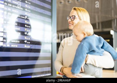 Mutter reist mit Kind, hält seinen kleinen Jungen am Flughafenterminal, überprüft Flugplan, wartet auf das Einsteigen in ein Flugzeug. Das Konzept „Reisen mit Kindern“ Stockfoto