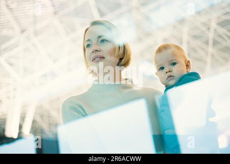 Die nachdenkliche junge Mutter schaute durch das Fenster und hielt sein kleines Kind fest, während sie darauf wartete, an den Abflugsteigen des Flughafenterminals an Bord eines Flugzeugs zu gehen. Das „Reise mit Baby“-Konzept Stockfoto