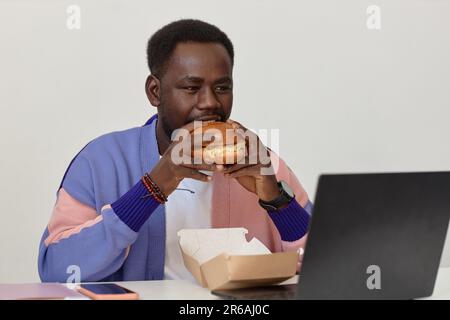 Porträt eines jungen Schwarzen, der Burger zum Mitnehmen am Arbeitsplatz isst, an der weißen Wand, Kopierraum Stockfoto