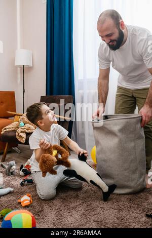 Vater und kleiner Junge räumen zusammen Spielzeuge auf, legen Textilkorb in die Wohnzimmereinrichtung, viel Spaß mit der Familie Stockfoto