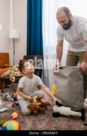 Vater und kleiner Junge räumen zusammen Spielzeuge auf, legen Textilkorb in die Wohnzimmereinrichtung, viel Spaß mit der Familie Stockfoto