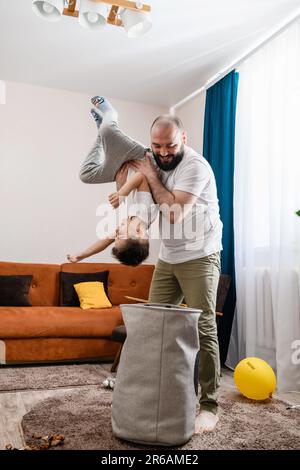 Vater und kleiner Junge räumen zusammen Spielzeuge auf, legen Textilkorb in den Wohnzimmerraum, Junge steht auf dem Kopf Stockfoto