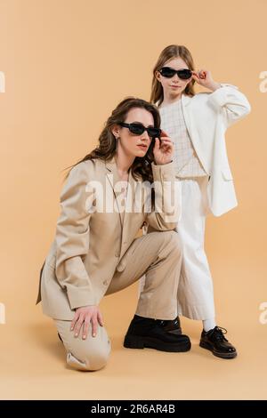 Moderne Familie, stilvolle Mutter und Tochter in Anzügen und Sonnenbrillen, Geschäftsfrau, die in der Nähe des Mädchens auf beigefarbenem Hintergrund sitzt, modische Outfits, formelles A Stockfoto