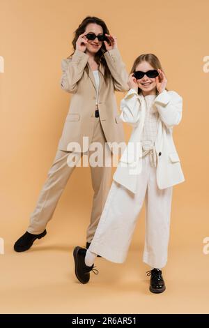 Stilvolle Mutter und Tochter in Sonnenbrille, glückliche Geschäftsfrau und Mädchen in beigefarbenen Anzügen, modische Outfits, formell AT Stockfoto