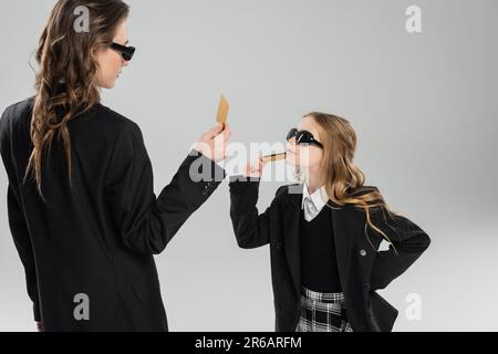 Mutter und Tochter in Sonnenbrille, Geschäftsfrau im Anzug und Schulmädchen in Uniform, die Kreditkarten hält und sich auf grauem Hintergrund ansieht, Stockfoto