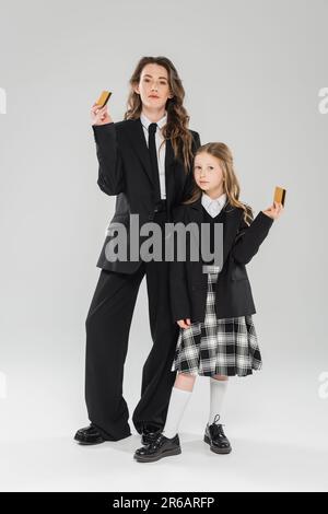 Trendige Mutter und Tochter, Geschäftsfrau im Anzug und Schulmädchen in Uniform mit Kreditkarten auf grauem Hintergrund, moderne Elternschaft, Finanzerziehung Stockfoto