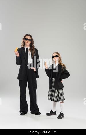 Mutter und Tochter in Sonnenbrille, Geschäftsfrau im Anzug und Schulmädchen in Uniform mit Kreditkarten auf grauem Hintergrund, moderne Erziehung, Financia Stockfoto