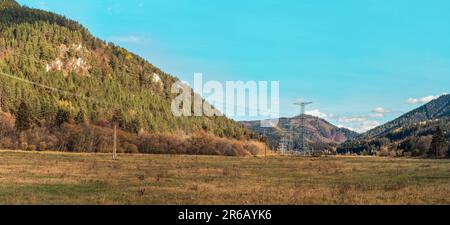 Große Metall-Strommasten unter Stromleitungen, auf dem Land gebaut mit Herbstgras und waldbedeckten Hügeln, blauer Himmel Hintergrund Stockfoto