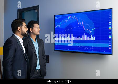 Indische Geschäftskollegen schauen sich Aktien online im Büro an. Börsenmakler, die Indizes auf dem großen Computerbildschirm ansehen. Finanzkennzeichen Stockfoto