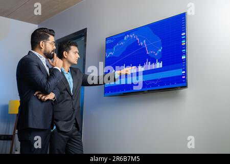 Indische Geschäftsleute, die online im Büro über Aktien handeln und diskutieren. Börsenmakler, die Indizes auf dem großen Computerbildschirm ansehen. Stockfoto