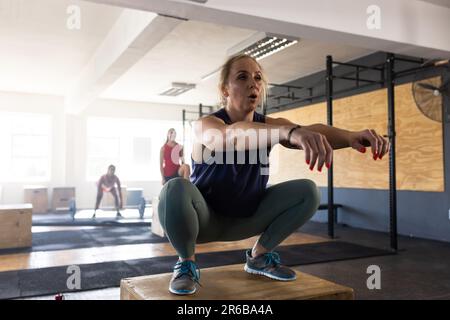 Volle Länge einer engagierten weißen Frau, die Squats auf hölzernen Plyoboxen im Fitnessclub praktiziert Stockfoto