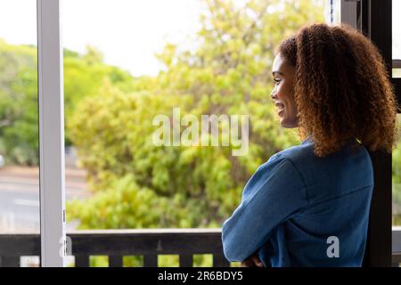 Glückliche, birassische Frau mit lockigem Haar auf dem Balkon, die Bäume ansieht und lächelt, mit Kopierraum Stockfoto