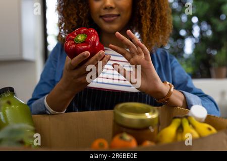 Glückliche birassische Frau, die Lebensmittel auspackt, aus einer Schachtel in der Küche mit rotem Pfeffer einkauft Stockfoto