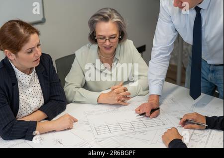 Ein weißer Mann steht und ändert eine Zeichnung, drei Kollegen sitzen an einem Tisch und hören ihm zu. Stockfoto