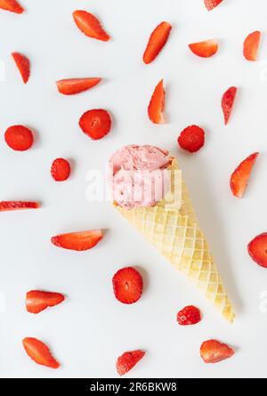 Erdbeereis in einem Waffelbecher in Form einer Schale, Eis liegt auf einem weißen Tisch zusammen mit Scheiben frischer Erdbeeren. Vertikales Foto Stockfoto