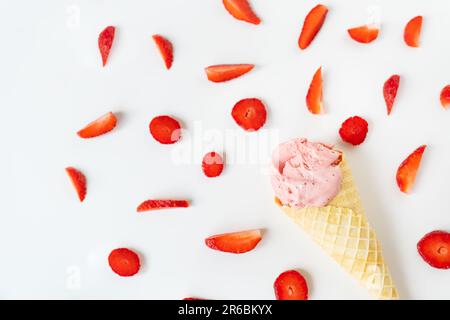 Erdbeereis in einem Waffelbecher in Form einer Schale, Eis liegt auf einem weißen Tisch zusammen mit Scheiben frischer Erdbeeren Stockfoto