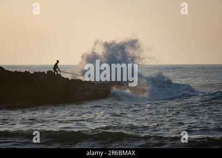 Ein Silhouettenfischer, der auf einem felsigen Atlantik-Felsvorsprung steht, mit seiner Angelrute, die auf die schlagenden Wellen in Oualidia, Marokko, gerichtet ist Stockfoto