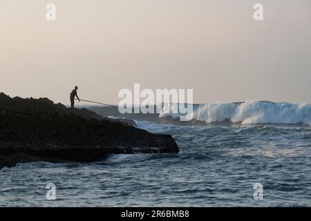 Ein Silhouettenfischer, der auf einem felsigen Atlantik-Felsvorsprung steht, mit seiner Angelrute, die auf die schlagenden Wellen in Oualidia, Marokko, gerichtet ist Stockfoto