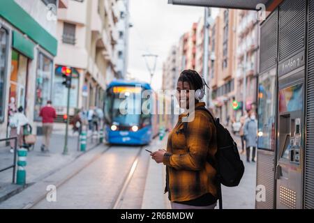 Junger Mann, der Musik hört, während er auf die Straßenbahn an der Haltestelle wartet. Konzept: Öffentliche Verkehrsmittel, Lifestyle, Outdoor Stockfoto