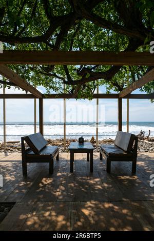 Elegantes Set aus Tisch und Stuhl auf einem hölzernen Dock am Pazifik unter dem Schatten einer grünen Pflanze Stockfoto