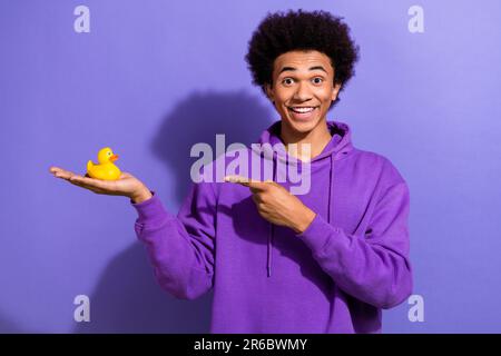 Foto eines optimistisch beeindruckten Mannes mit einem violetten Hoodie im Afro-Stil, der auf dem Entenspielzeug isoliert auf violettem Hintergrund angezeigt wird Stockfoto