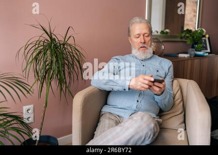 Porträt eines verblüfften, grauhaarigen, reifen Mannes, der sein Smartphone mit der mobilen Online-App hält und mit offenem Mund auf dem Sessel auf den Bildschirm schaut Stockfoto