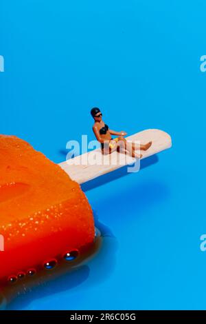 Eine Miniaturfrau, die einen schwarzen Bikini trägt, wird gleich Sonnencreme auf ihre Beine auftragen und sitzt auf einem schmelzenden orangefarbenen Eis am Stiel auf blauem Hintergrund Stockfoto