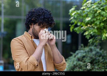 Ein junger hispanic steht auf der Straße und wischt sich die Nase mit einem Taschentuch. Er hat eine laufende Nase, Grippe, leidet an saisonalen Allergien. Stockfoto