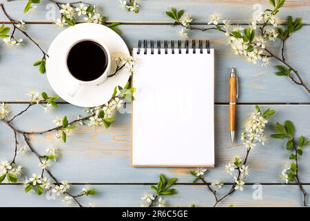 Künstlerische Komposition mit geöffnetem Notizblock und Stift, Tasse schwarzen Kaffee, Zweig weißer Kirschblüten auf blauem Holztisch. Festlicher Büroschreibtisch Stockfoto