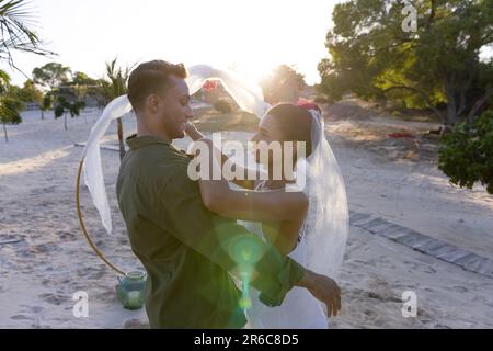 Blick von der Seite auf ein frisch verheiratetes Paar, das während der Hochzeitszeremonie am Strand bei Sonnenuntergang tanzt Stockfoto
