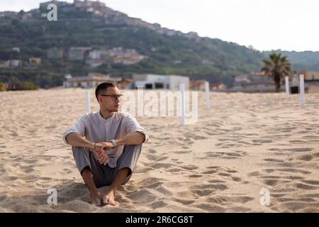Ein junger weißer Mann in einem kragenlosen Hemd und einer Kleiderhose, sitzt auf dem Sand und schaut zur Seite Stockfoto