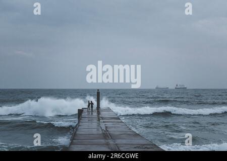 Silhouette von 2 jungen Surfern auf einer Betonplattform mit brechenden Wellen an stürmischen Tagen Stockfoto