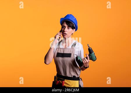 Überraschter Baumeister, der telefonisch spricht und eine Bohrpistole in der Hand hält, mit Smartphone im Studio. Weibliche Auftragnehmerin, die vor der Renovierung des Projekts per Ferngespräch am Handy teilnimmt. Stockfoto