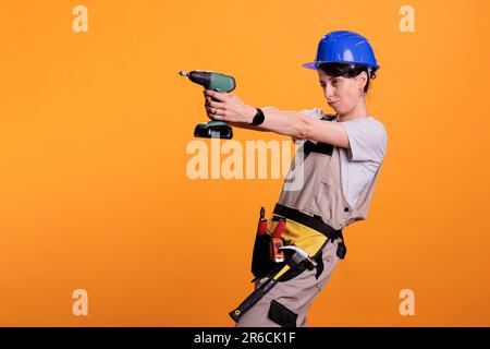 Eine professionelle Frau, die eine schnurlose Bohrpistole hält und Spaß hat, sich albern benimmt. Eine Baufirma trägt Schutzhelm und Uniform, macht sich lustig mit einer renovierten Nagelpistole. Stockfoto