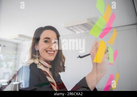 Eine glückliche Geschäftsfrau schreibt mit einem Stift auf den Strichzetteln, die an der Glaswand hängen Stockfoto