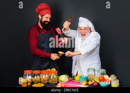 Zwei Köche in Uniform kämpfen in der Küche. Professionelles kulinarisches Angebot. Die Küche ist super. Kochwettbewerb. Kulinarische Schlacht in der Küche. Bärtige Köche auf Stockfoto