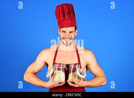 Ein lächelnder Koch mit Kochmütze und Schürze hält Gläser mit Grobgrieß und Müsli. Gesundes Essen Kochen. Lebensmittelzubereitung. Gesunde Ernährung. Professionell Stockfoto