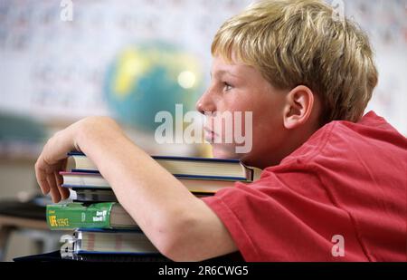 Ein abgenutzter Junge in der Schule, der im Unterricht nicht aufpasst, sich auf einen großen Stapel Bücher lehnt Stockfoto