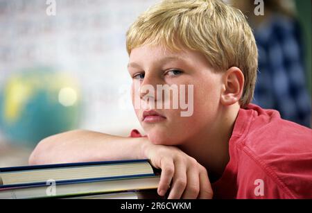 Ein abgenutzter Junge in der Schule, der im Unterricht nicht aufpasst, sich auf einen großen Stapel Bücher lehnt Stockfoto