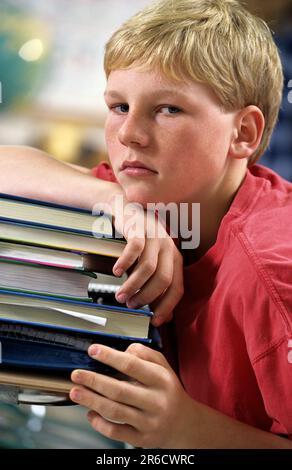 Porträt eines abgenutzten Jungen in der Schule, der im Unterricht nicht aufpasst, sich auf einen großen Stapel Bücher lehnt Stockfoto