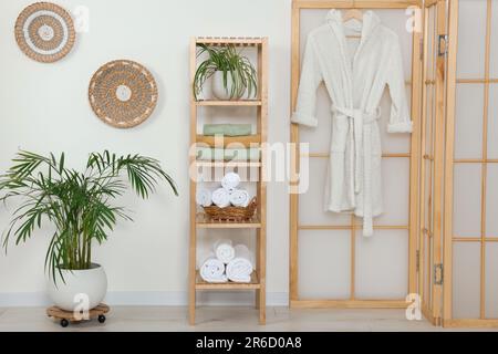 Weiche gefaltete Handtücher auf Holzregal und Bademantel nahe der weißen Wand Stockfoto